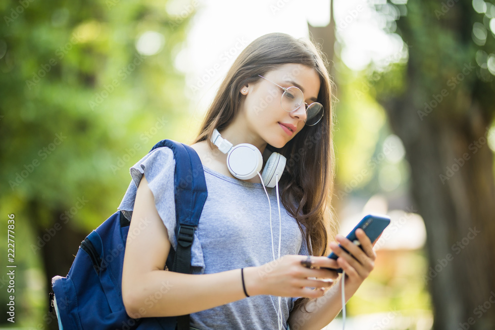 Smiling young girl student with backpack holding mobile phone with earphones, walking at the park