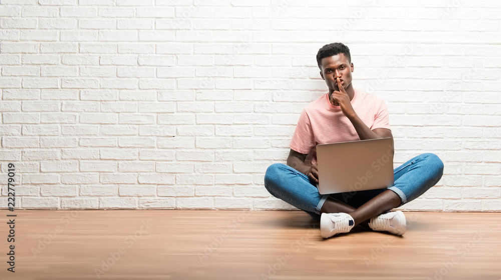 Afro american man sitting on the floor with his laptop showing a sign of closing mouth and silence gesture