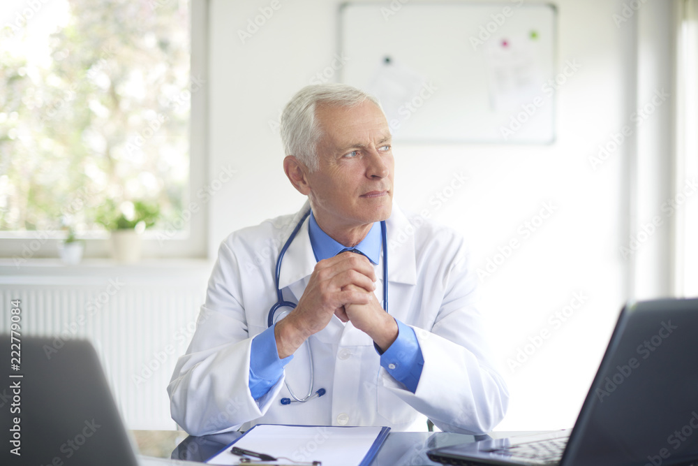 Thinking male doctor sitting in at desk behind computers in the consulting room.