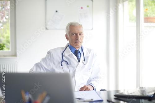 Thinking male doctor sitting in at desk behind computers in the consulting room.