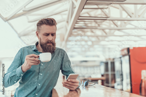 Portrait of concentrated bearded man drinking cup of coffee while typing in mobile indoor photo