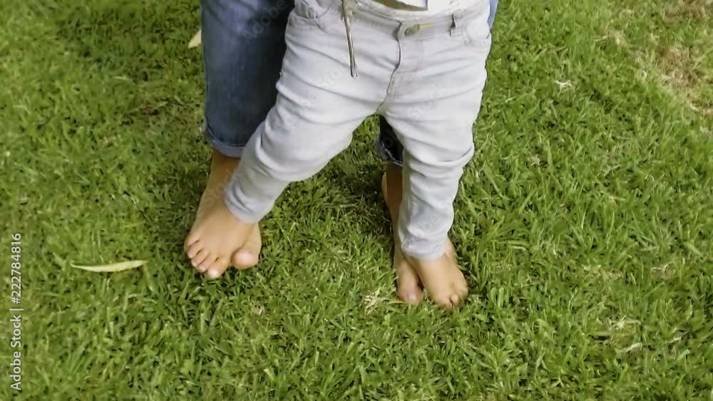 Barefoot parent teaching son to walk on grass in slow motion. Partial view of mom holding her little boy on legs and walking together. Parenting concept Barefoot parent teaching son to walk on grass in slow motion. Partial view of mom holding her little boy on legs and walking together. Parenting concept