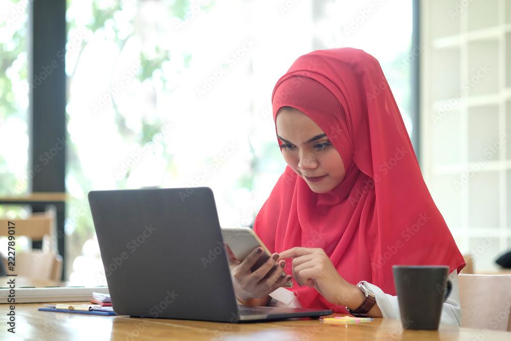 Attractive young Asian wearing red hijab using her mobile smartphone at workspace desk. Attractive young Asian wearing red hijab using her mobile smartphone at workspace desk.