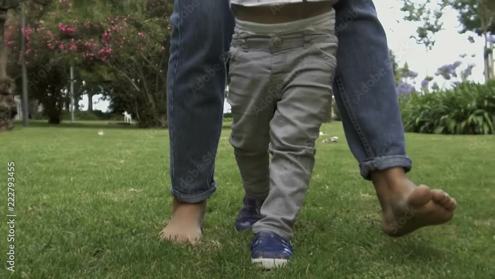 Mother with little son walking on lawn. Barefoot mom teaching cute boy to walk in in slow motion. Low section. Baby boy learning to walk with mother help. Childhood concept Mother with little son walking on lawn. Barefoot mom teaching cute boy to walk in in slow motion. Low section. Baby boy learning to walk with mother help. Childhood concept