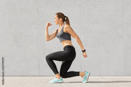 Indoor shot of self determined woman with pony tail, stands on one knee, warms up before cardio training, wears tanktop, leggings, sneakes, goes in for sport regularly, isolated over grey background
