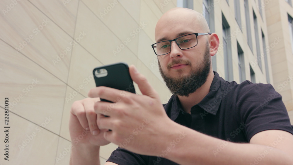 A man with a beard wearing black glasses uses a phone sitting in front of a large building. Close-up shot. Soft focus A man with a beard wearing black glasses uses a phone sitting in front of a large building. Close-up shot. Soft focus