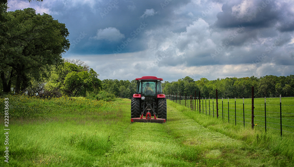 Fototapeta premium Farming: Large red tractor mowing green farmers pasture along barbwire fence