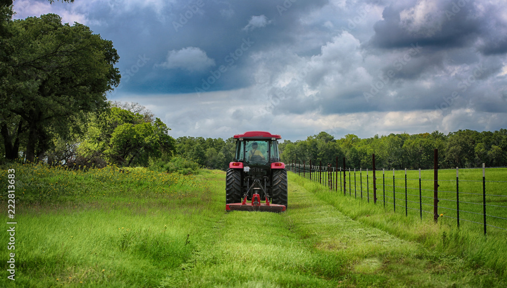 Obraz premium Farming: Large red tractor mowing green farmers pasture along barbwire fence