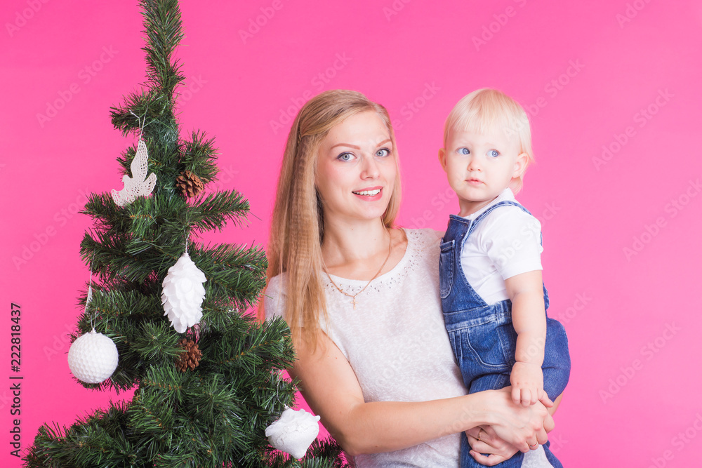 Holidays, family and christmas concept - young mom with her little daughter near christmas tree on pink background Holidays, family and christmas concept - young mom with her little daughter near christmas tree on pink background