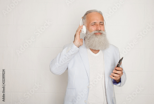 Portrait of unshaved male pensioner 60s with gray hair holding cellphone and listening to music via wireless headphones isolated over white background
