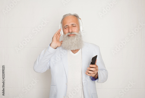 Portrait of unshaved male pensioner 60s with gray hair holding cellphone and listening to music via wireless headphones isolated over white background