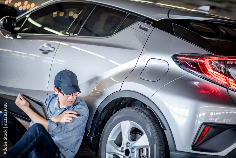 Young Asian auto mechanic holding digital tablet checking car wheel in auto service garage. Mechanical maintenance engineer working in automotive industry. Automobile servicing and repair concept