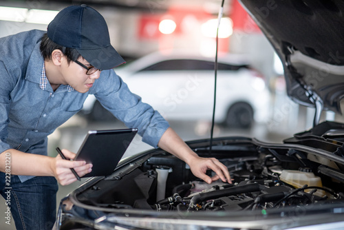 Asian auto mechanic holding digital tablet checking car engine under the hood in auto service garage. Mechanical maintenance engineer working in automotive industry. Automobile servicing and repair