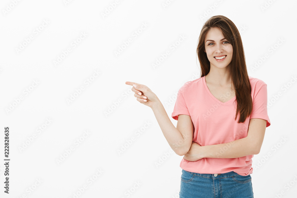 Portrait of beautiful brunette in pink t-shirt smiling joyfully while pointing left with index finger as if inviting to get inside or suggesting visit new store, posing over gray background Portrait of beautiful brunette in pink t-shirt smiling joyfully while pointing left with index finger as if inviting to get inside or suggesting visit new store, posing over gray background