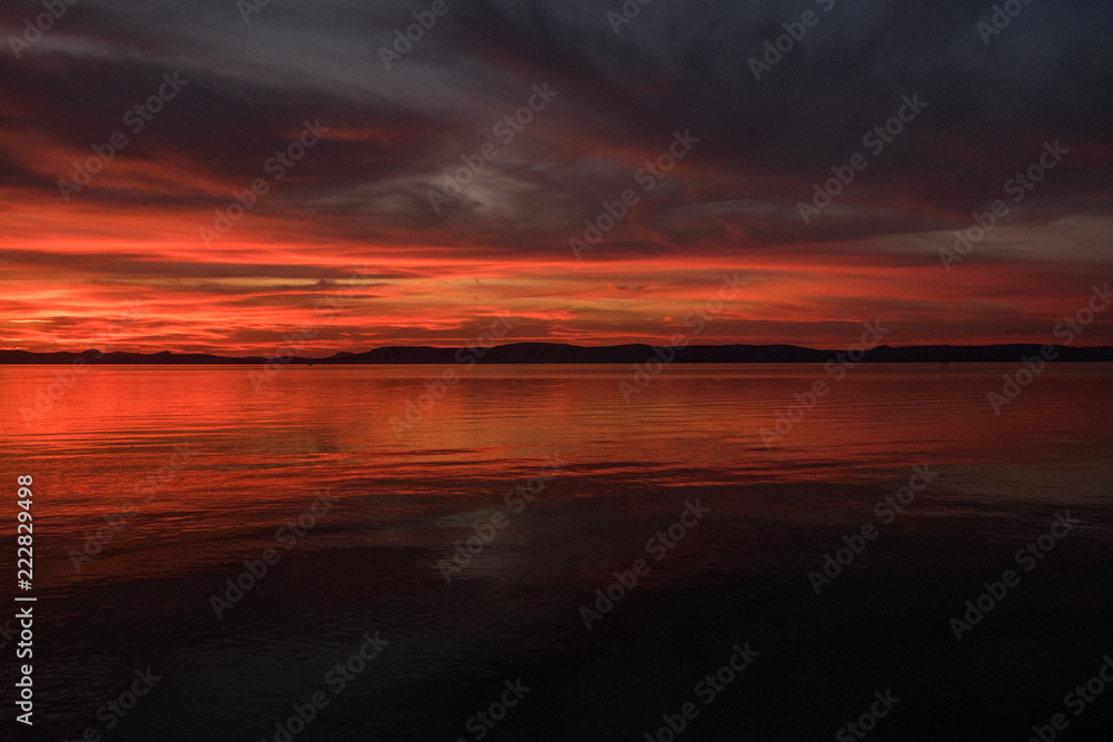 red and hot sunset at Balaton lake - clouds, thin waves on water and hills in background - dark tones wide angle