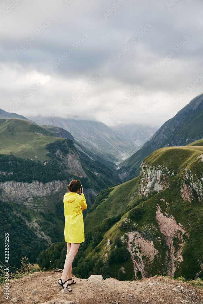Fototapeta premium A young Girl in a yellow raincoat photographs the mountains. Georgia. Summer. August. Girl making a photo shoot of mountain. 