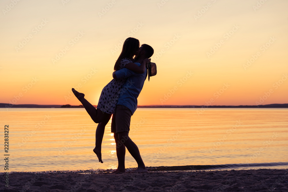 Silhouette of happy couple on beach at sunset, man taking the girl in his arms.