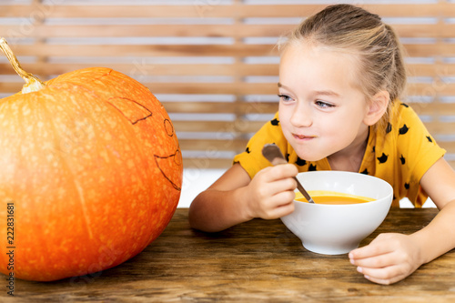 Cute little girl eating pumpkin soup and looking at a large Halloween pumpkin, with vicious face expression. Halloween conceptual background. photo