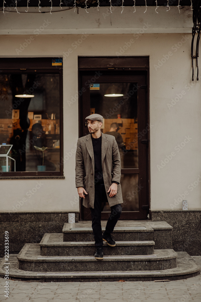 City life. Filtered effect. Stylish young American man standing on the street. Businessman wearing a grey fashionable trench coat and hat. Guy lookin outside.