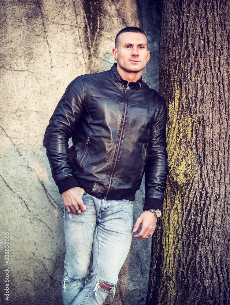 One handsome young man in urban setting leaning against tree, standing, wearing black leather jacket and jeans