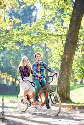 Young active happy traveler couple, handsome bearded man and attractive blond woman smiling and cycling together tandem double bike along path in the morning in autumn park under tall trees.