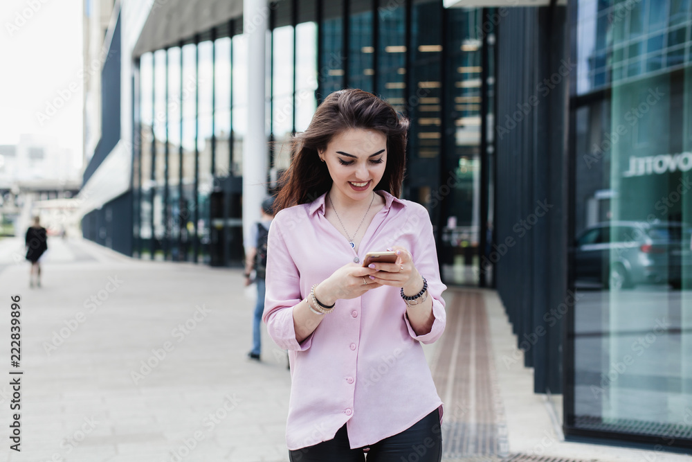Happy pretty brunette girl using mobile phone near office, beautiful woman browsing phone and doing online shopping in a mobile app while smiling walking near shopping mall. Online communication. Happy pretty brunette girl using mobile phone near office, beautiful woman browsing phone and doing online shopping in a mobile app while smiling walking near shopping mall. Online communication.