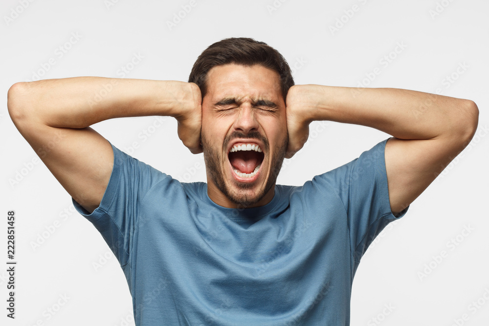 Young man in blue t-shirt, screaming with closed eyes, stressed by noise, closing ears with both hands - obrazy, fototapety, plakaty 