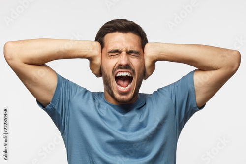 Young man in blue t-shirt, screaming with closed eyes, stressed by noise, closing ears with both hands