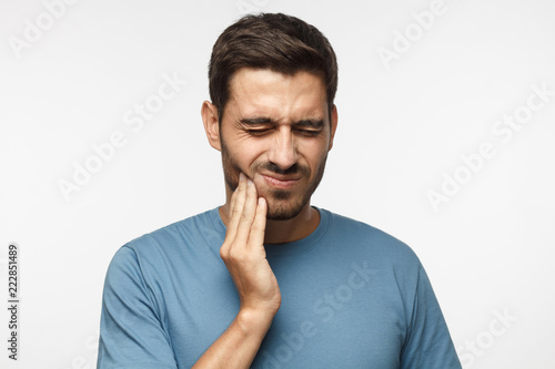 Tooth ache concept. Indoor shot of young male feeling pain, holding his cheek with hand, suffering from bad toothache, looking at camera with painful expression photo
