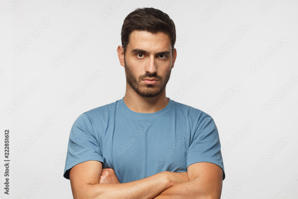 Obraz premium Young man in blue t-shirts tanding with arms crossed and serious concentrated face at camera, looking aggressive, isolated on gray background