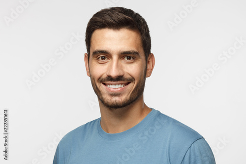 Headshot of young handsome european caucasian man isolated on gray background. wearing casual blue t-shirt, smiling happily and friendly at camera, looking confident and relaxed