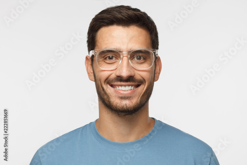 Close up shot of smiling attractive man in blue t-shirt and trendy trasparent eyeglasses isolated on gray background © Damir Khabirov
