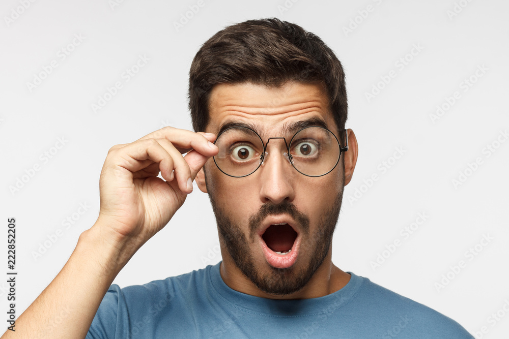Young man in blue t-shirt shouting oh my god with open mouth, holding round glasses, surprised by low price and sales Young man in blue t-shirt shouting oh my god with open mouth, holding round glasses, surprised by low price and sales