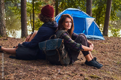 Young hipster couple hiking in the forest and sitting back to back while warming near a campfire at camp.