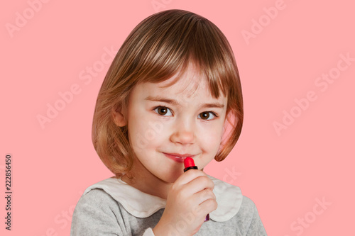 Portrait of smiling little baby girl paints lips with lipstick isolated on a pink background.