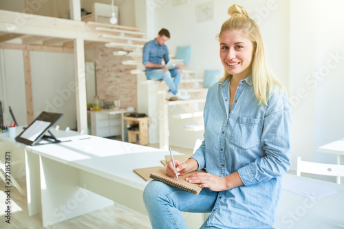 Positive young blonde female in denm shirt and blue jeans sketching in notepad while sitting on desk in studio of design photo