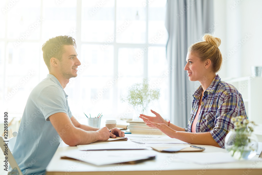 Two young confident managers in casualwear sitting by desk in front of each other and discussing ideas Two young confident managers in casualwear sitting by desk in front of each other and discussing ideas