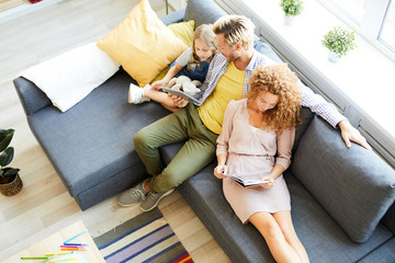 Overview of relaxed family of three sitting on sofa, reading and watching online movie on weekend