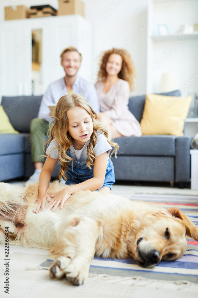 Litle girl cuddling fluffy dog lying on the floor of living-room with her parents sitting on background Litle girl cuddling fluffy dog lying on the floor of living-room with her parents sitting on background