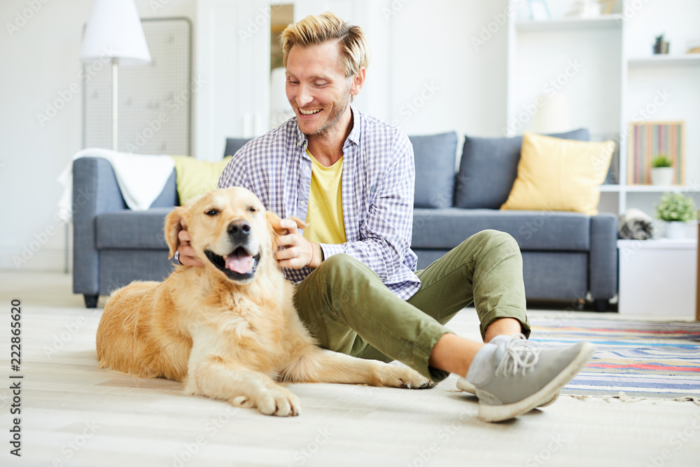Cheerful young man in casualwear relaxing at home and playing with friendly purebred golden labrador