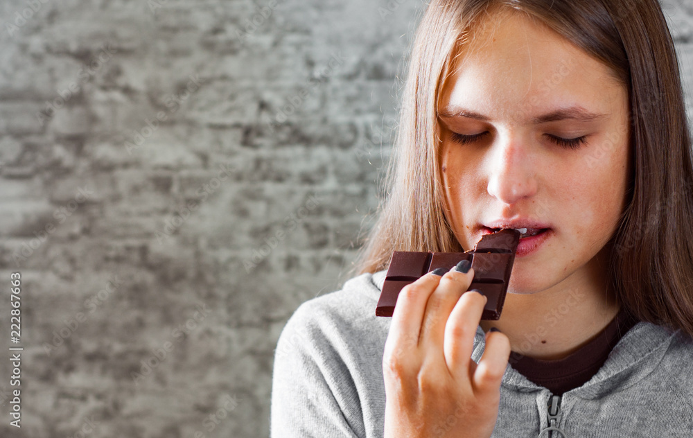 portrait of young teenager brunette girl with long hair eating chocolate on gray wall background