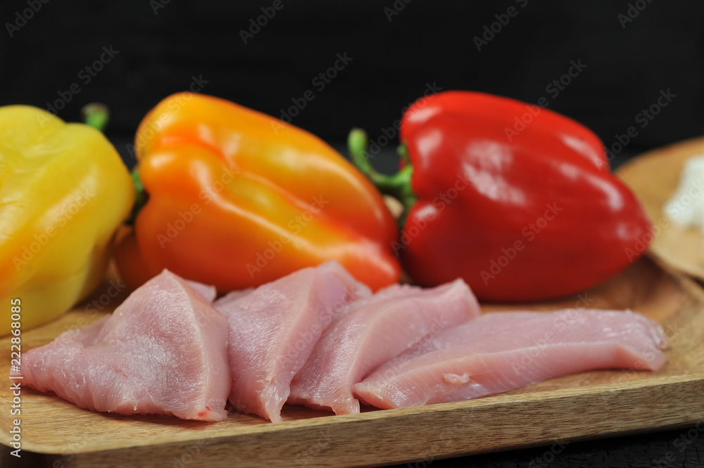 Yellow, orange, red bell peppers on a wooden plate. Next to the peppers are a few cheese steaks from turkey meat. Black background. Close-up. Macro photography.