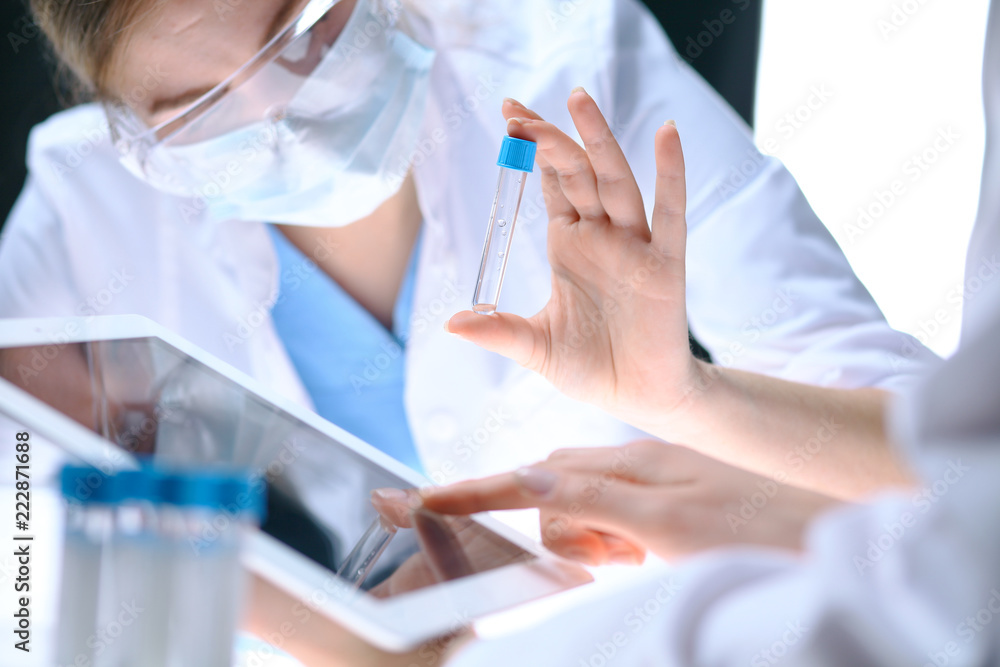 Closeup of scientific research team with clear solution in laboratory. Blonde female chemist holds test tube of glass while her colleague checks results with tablet pc. Blood test, medicine or Closeup of scientific research team with clear solution in laboratory. Blonde female chemist holds test tube of glass while her colleague checks results with tablet pc. Blood test, medicine or