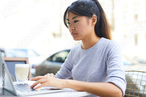 Young Asian student in grey pullover sitting by table in front of laptop, having drink and networking in cafe Free Download Young Asian student in grey pullover sitting by table in front of laptop, having drink and networking in cafe - GFXLoad