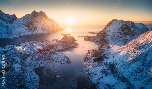 Fototapeta samoprzylepna Widok z lotu ptaka piękny fjord przy zmierzchem w Lofoten wyspach, Norwegia. Zimowy pejzaż z ośnieżonymi górami, błękitnym morzem i niebem ze słońcem o zmierzchu. Widok z góry skał na śniegu, drodze, wsi. Wybrzeże północne