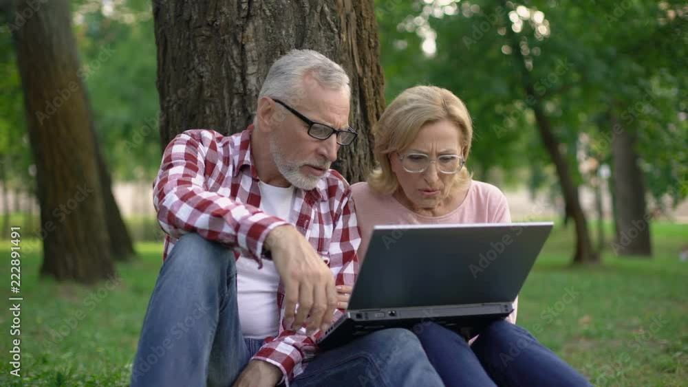 Happy retired man and woman sitting on grass and watching movie on laptop pc