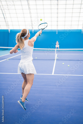 Back view action portrait of female tennis player swinging racket in jump during training in indoor court, copy space