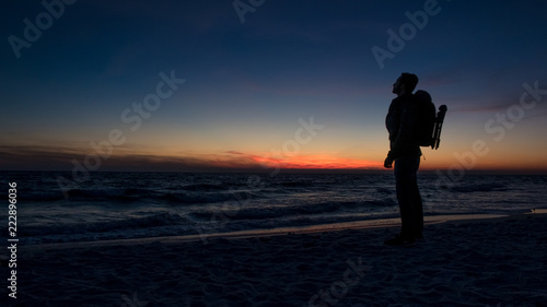 Guy stands in front of dramatic sunset over the ocean on a beach covered with waves.