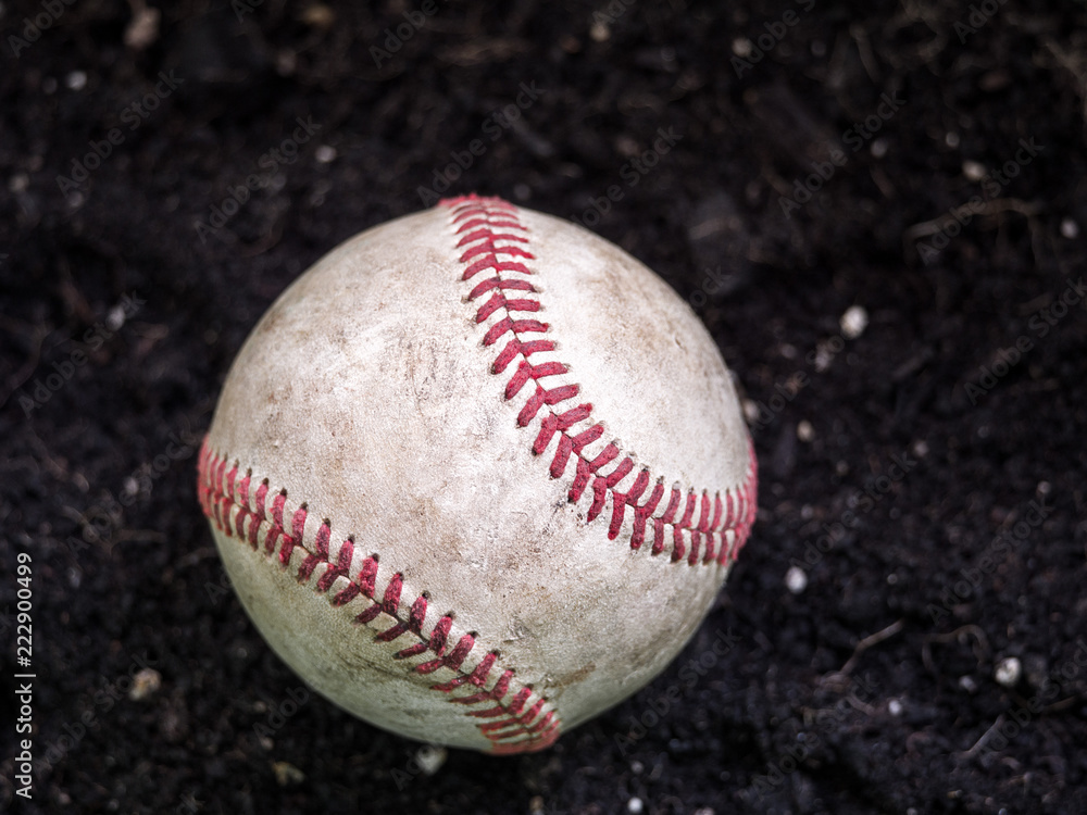 Close up sports background image of an old used weathered leather baseball laying in a pile of brown dirt showing intricate detailing and red laces.