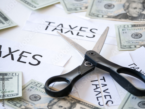 Close up photograph of a scissors cutting a paper that has taxes typed on it with 20 dollar US money representative of government tax cuts and saving money. photo